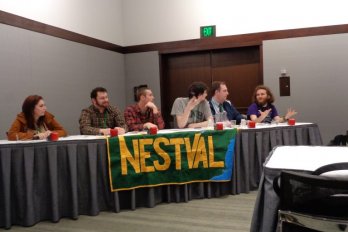 6 students seated, panel style, at table in hotel conference room, facing camera, with large banner displaying letters 'NESTVAL' draped over front of table.