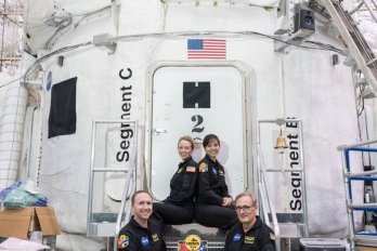 NASA crewmembers, in flight uniform, seated in front of NASA-white space capsule housed in aircraft hangar