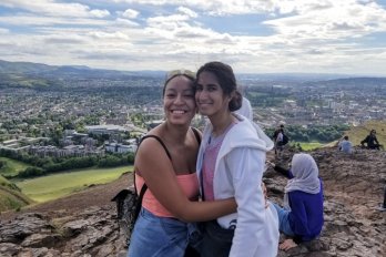 Raquel Bryant and Julie Beck facing camera in half-embrace atop rock outcrop in front of green pastoral scenery surrounding Edinburgh