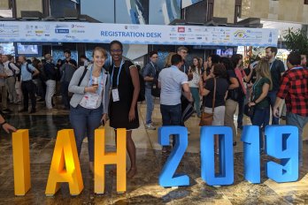 Candid photo of Sarah McKnight (left) and Marsha Allen (right) in front of crowd at a conference center, standing behind knee-high orange and blue block letters saying "IAH 2019"