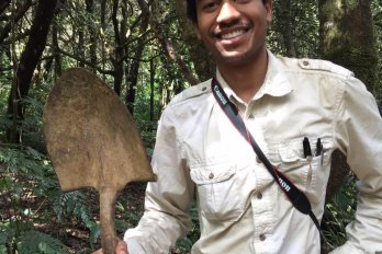 Portrait of Dr. Justin Richardson in field clothing in a forest, smiling at camera, holding shoveled with one hand, blade up.