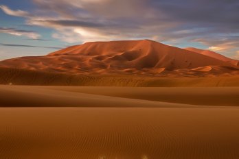 Brown hill in a sandy desert landscape under partly cloudy blue sky near the end of day.