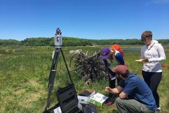 Hydrogeology students standing in a field with survey and geophysical equipment, adjacent to marshland