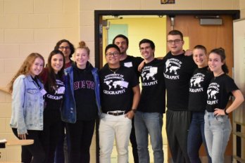 Members of U-Mass Geography club assembled for group photo in front of doorway in lecture all, smiling at camera and wearing Geography Club t-shirts