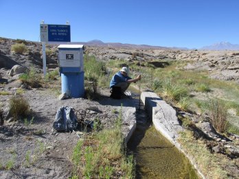 Dr. David Boutt, crouched by small, short, concrete lined water channel in the Salar de Atacama, surrounded by desert landscape with brush and brown snow-capped mountains and clear blue sky.