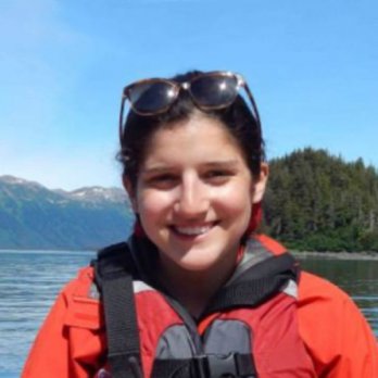 Portrait of Kaitlyn Suarez in front of mountain lake with snow-capped peaks in background. Kaitlyn is wearaing safety-orange foul-weather gear and is smiling at camera