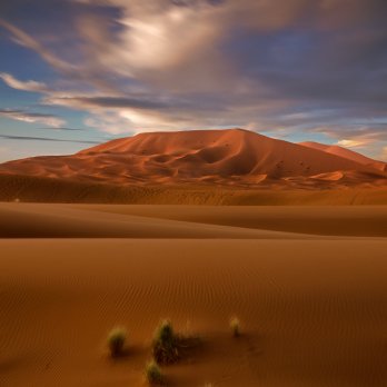 Brown hill in a sandy desert landscape under partly cloudy blue sky near the end of day.