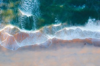 Aerial view of surf zone of beach, with green-blue water dominating top half of photograph, white foamy surf in middle, and pink-yellow beach at bottom.