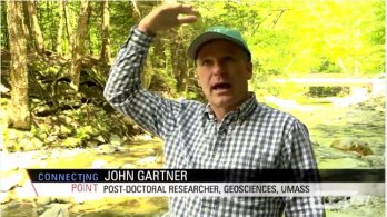 Screen capture of post-doc John Gartner speaking to camera in front of Chicklea River in Hawley, MA. John is gesturing to show height of floodwaters above his head.