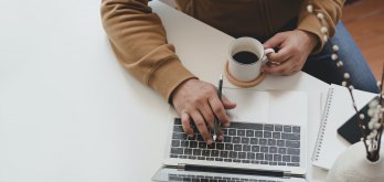 Hands and forearms of person using notebook computer at white table while drinking coffee and holding a pen.